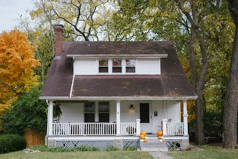 White clapboard cottage with a covered front porch and autumn trees